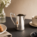 Vintage silver stainless steel pitcher on a dark stone table with breakfast setting.