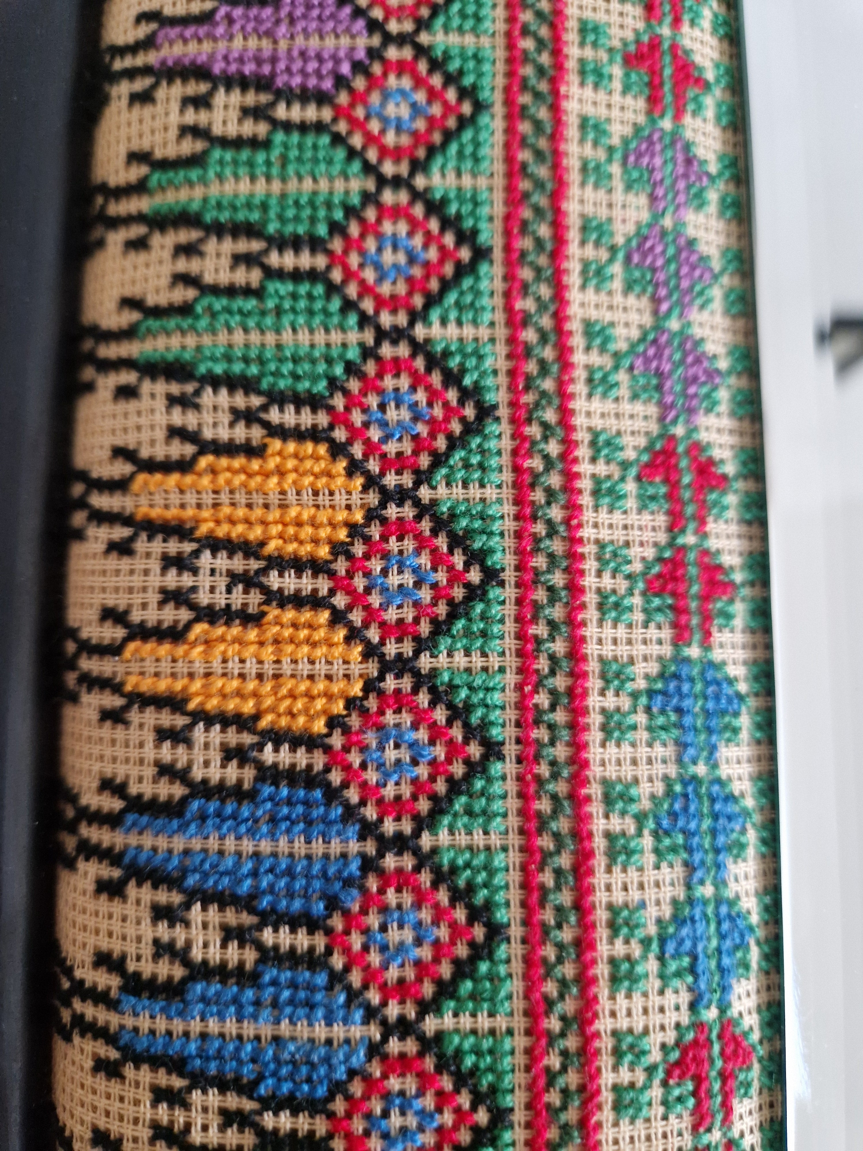 Close-up of hand cross-stitch embroidery showing geometric diamond motifs, birds and tulips in red, green, blue and gold on linen