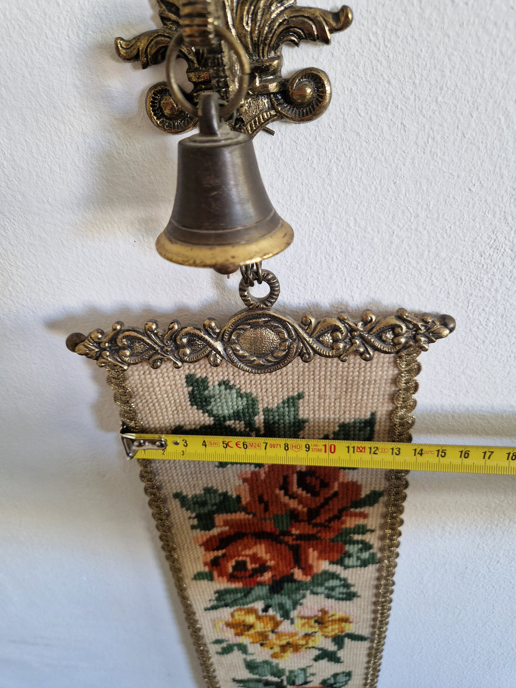 Close-up of top mount of antique bell pull showing Baroque brass finial, working bell on chain, and ornate scrollwork header with width measurement approx 15cm